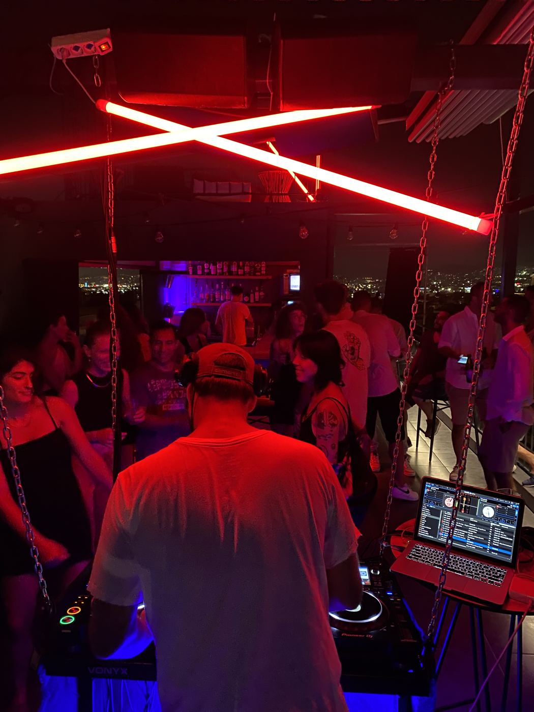 DJ deck under red neon X — rooftop crowd, Athens by night